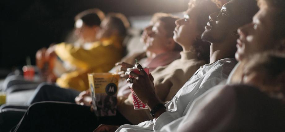 Audience members seated in a darkened movie theater, looking up at the screen while holding popcorn and drinks.
