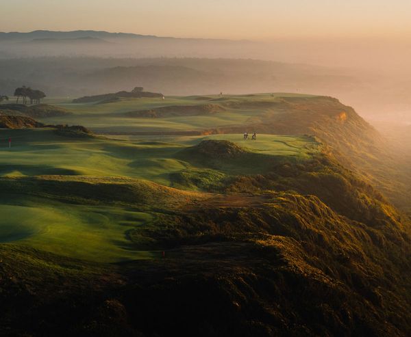 Aerial view of a coastal golf course at Bandon Dunes in Bandon, Oregon, showing rolling fairways, bunkers, and golfers on a cliffside green overlooking the Pacific ocean at sunset.