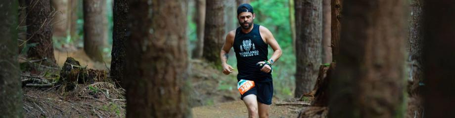 A man with a race bib runs a wooded trail wearing a Woodlands Running Co. shirt during Run Whiskey Run.
