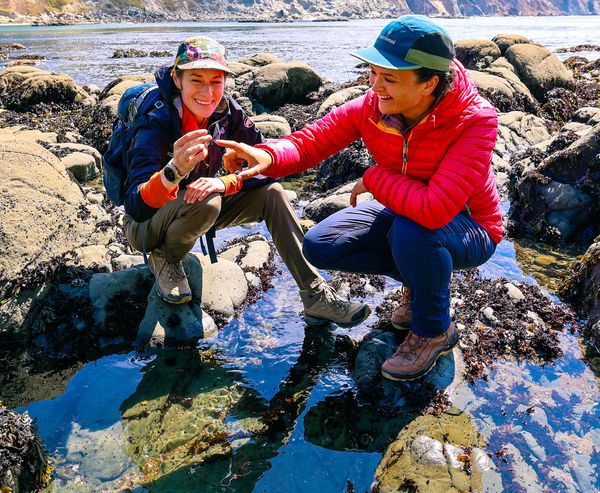 Two women crouched on rocks examining a small find together beside a clear tidepool, with a rugged coastline and ocean in the background.