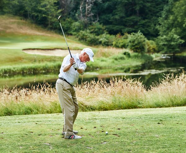 An older man in a white hat and polo swings a golf club on the tee box, with a pond and tall grasses behind him at Sunset Bay Golf Course.