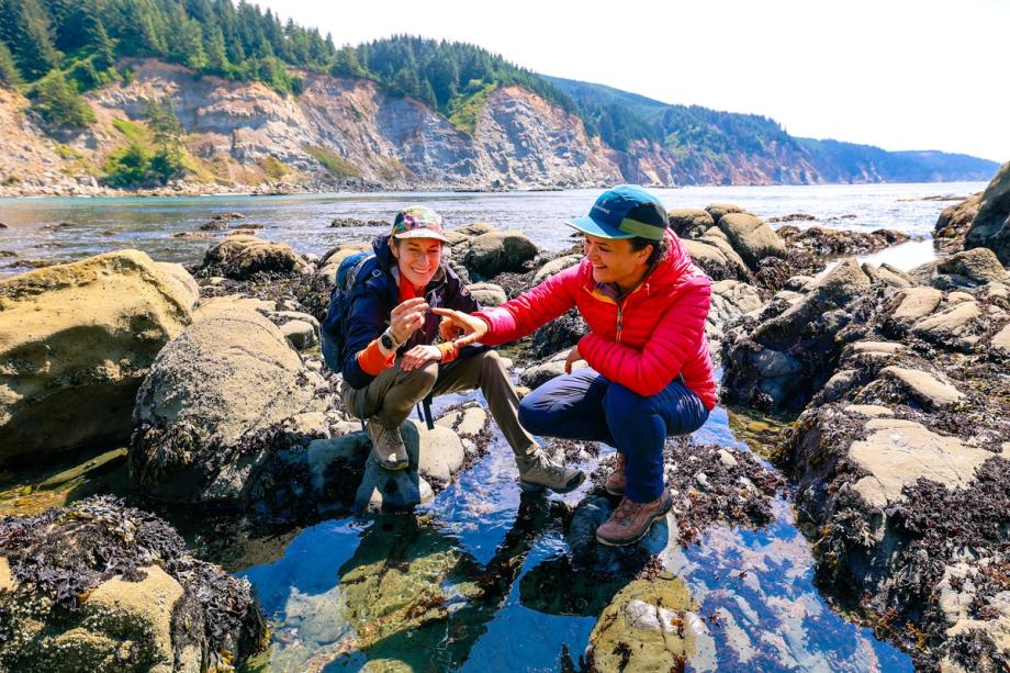 Two women in hiking gear crouch among tidepools on a rocky coastline, smiling as they examine a small find, with rugged cliffs and forested hills in the background.