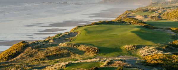 Golf green on a dune bluff at Bandon Dunes in Bandon, Oregon, overlooking a wide sandy beach and ocean waves at golden hour.