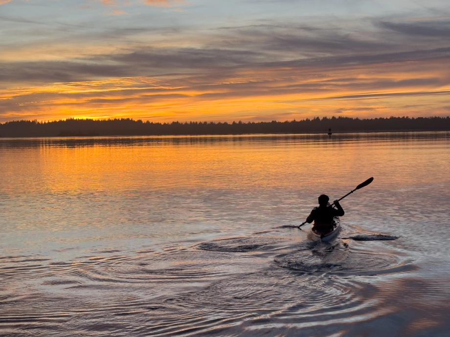 A silhouetted kayaker paddles across calm water toward a vivid orange and gold sunset, with a treeline reflected on the surface along the Oregon Coast.