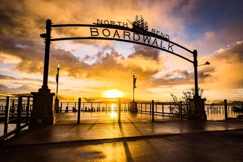 The arched entrance gate to the North Bend Boardwalk at sunrise, with golden light reflecting off Coos Bay and dramatic clouds filling the sky behind it.