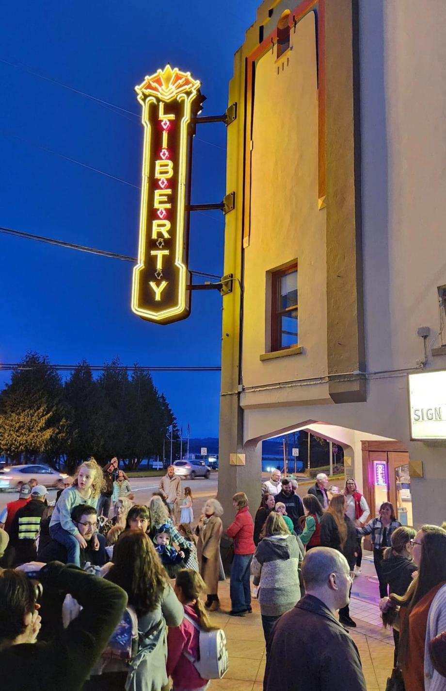 A crowd gathered at dusk beneath the illuminated yellow and red neon Liberty theater marquee sign mounted on a building facade.