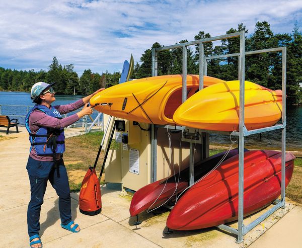 Woman lifting a yellow kayak from a self-service rental rack station beside a lake.