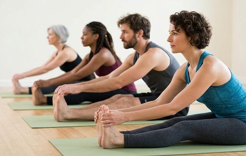 Four people performing a seated forward bend stretch in a yoga class, each on a green yoga mat.