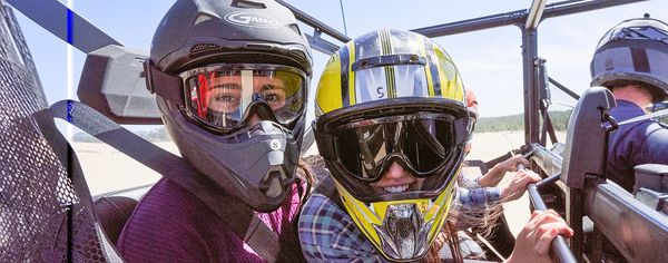 Two riders in helmets and goggles smile from inside a dune buggy, with a third rider visible behind them.