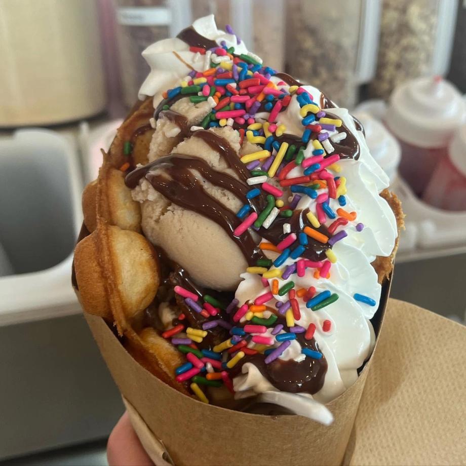 A photo of Scoops’ bubble waffle cone filled with ice cream, whipped cream, chocolate drizzle, and rainbow sprinkles, held in a paper wrap.