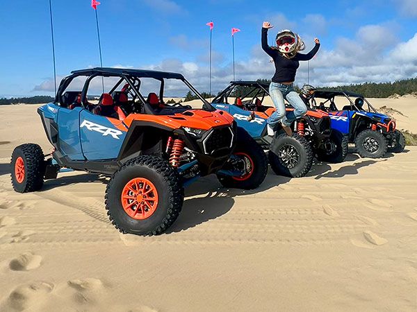 Here's the alt tag for this image: alt="A woman jumps for joy on top of a Polaris RZR dune buggy rental from Spinreel Dune Buggy and ATV Rentals, with a line of colorful ATVs parked on the sandy Oregon Dunes National Recreation Area.