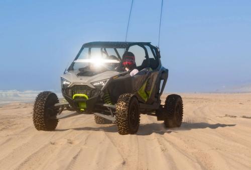 An off-road buggy with two goggled passengers drives across a foggy beach with ocean waves in the background.
