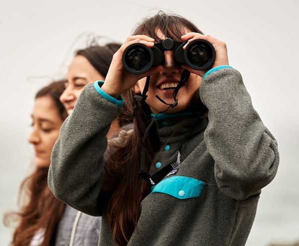 A smiling woman looks through binoculars while two friends stand behind her, all outdoors on an overcast day.