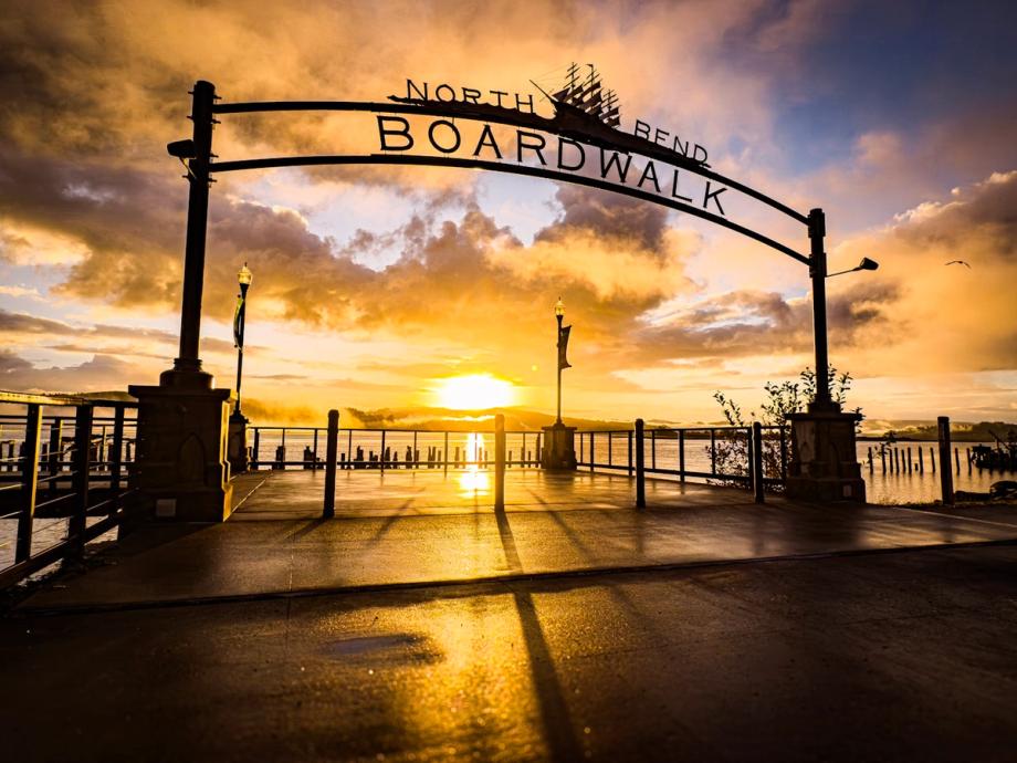 The North Bend Boardwalk archway silhouetted against a vivid golden sunrise over the water, with wet pavement reflecting the glowing sky.