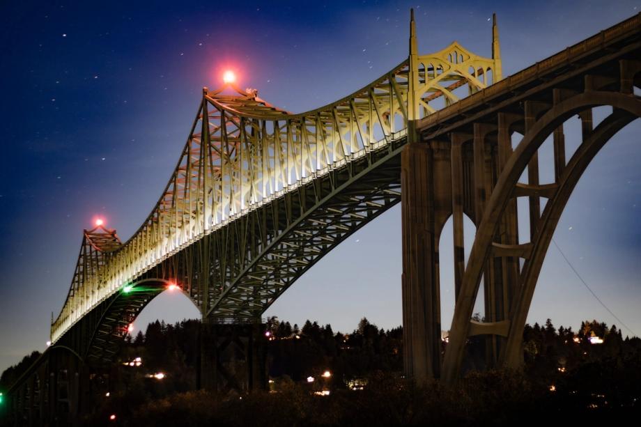 The Conde B. McCullough Memorial Bridge in North Bend, a steel arch truss bridge, lit up at night against a starry twilight sky with silhouetted trees below.
