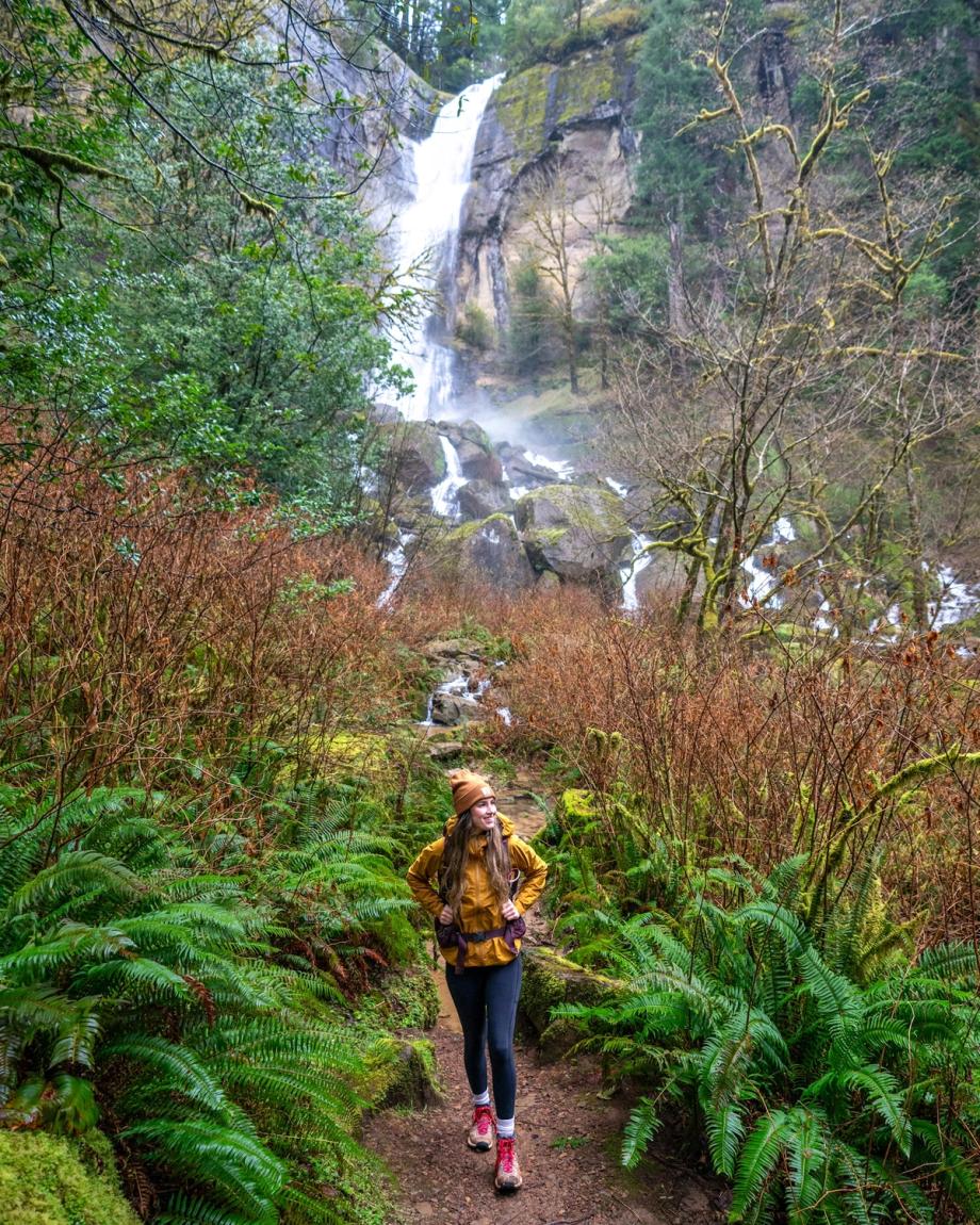 Hiker in yellow walks along a fern-lined trail in front of Golden Falls at Golden and Silver Falls Natural Area in Coos County, Oregon