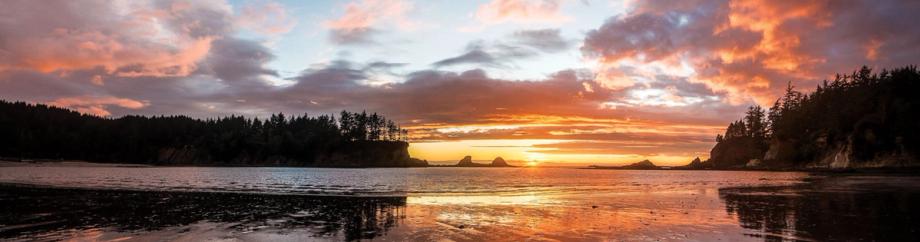 Panoramic sunset view from Sunset Bay State Park beach, with sea stacks silhouetted against a vivid orange and pink sky, reflected in the wet sand and calm tidal waters.