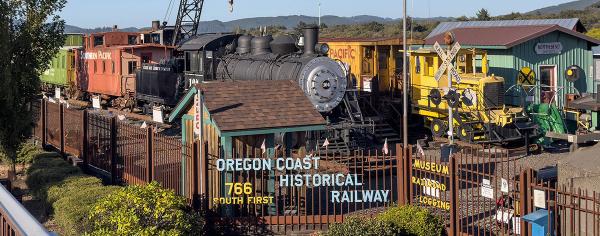 An aerial view of the Oregon Coast Historical Railway Museum in North Bend, featuring steam locomotive No. 104, a Southern Pacific caboose, vintage rail equipment, and the museum's entrance sign.