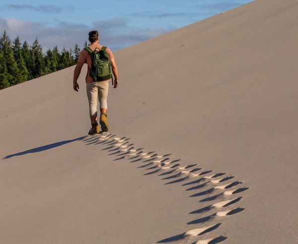 A man with a backpack walks up a large sand dune, leaving a trail of footprints behind him, with evergreen trees in the distance at Oregon Dunes National Recreation Area.
