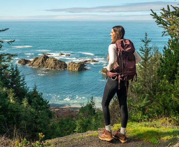A woman with a hiking backpack stands on a coastal overlook viewing rocky sea stacks and the ocean along the Sunset Trail.