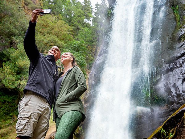 A couple takes a selfie in front of the tall, cascading Silver Falls waterfall surrounded by lush green trees and mossy rock walls