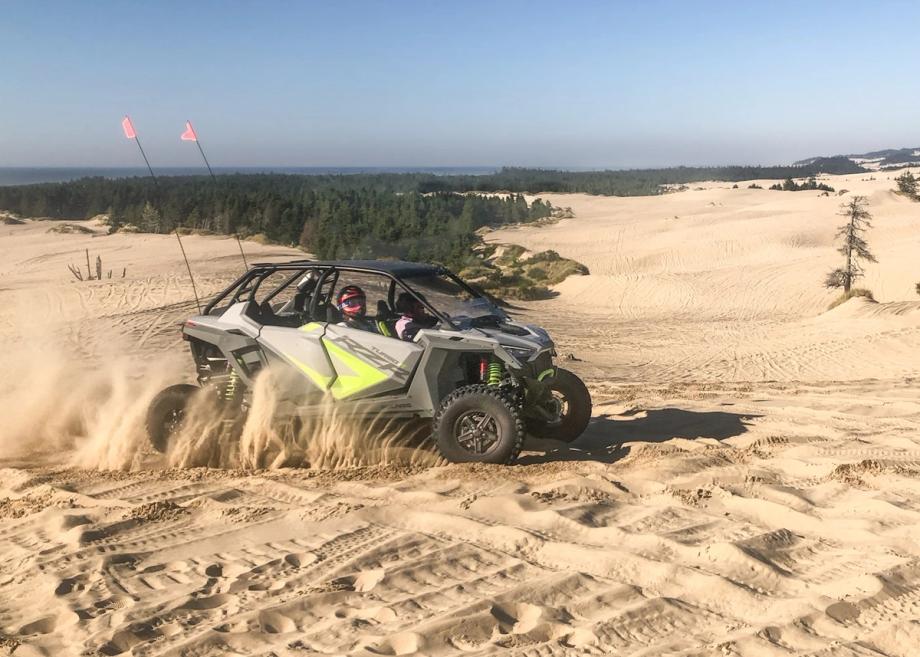 Silver and black Polaris RZR off-road vehicle with two helmeted riders kicking up sand while driving across desert dunes, with tall orange safety whip flags, a pine forest, and the ocean visible in the background under a clear blue sky.