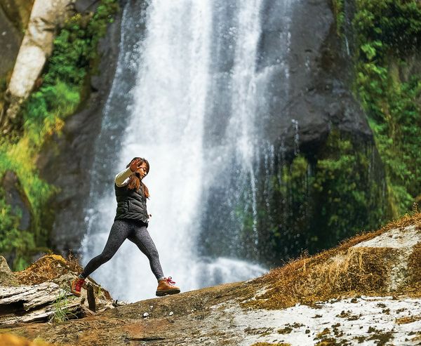 A woman in hiking boots crosses a rocky ledge near the base of a waterfall, holding back her hair as mist blows past.