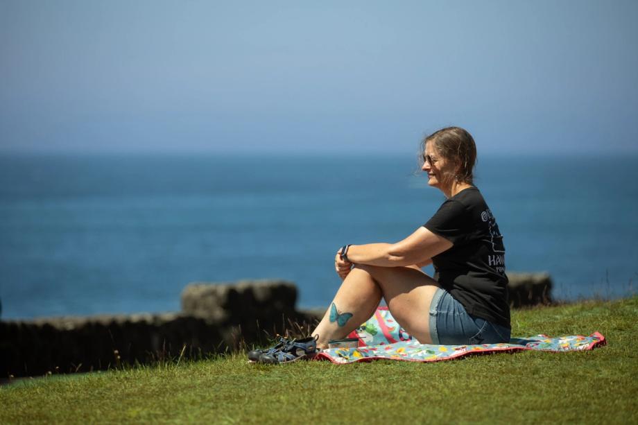 Woman in a black t-shirt and denim shorts sits on a picnic blanket on a grassy clifftop in Shore Acres State Park in Charleston, gazing out at the Pacific Ocean on a sunny day.