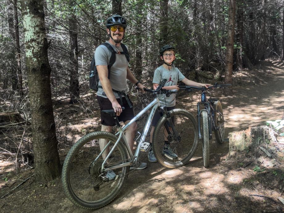 Man and boy in helmets stand smiling with their mountain bikes on a dirt trail in a wooded forest along the Whiskey Run Mountain Biking Trail.