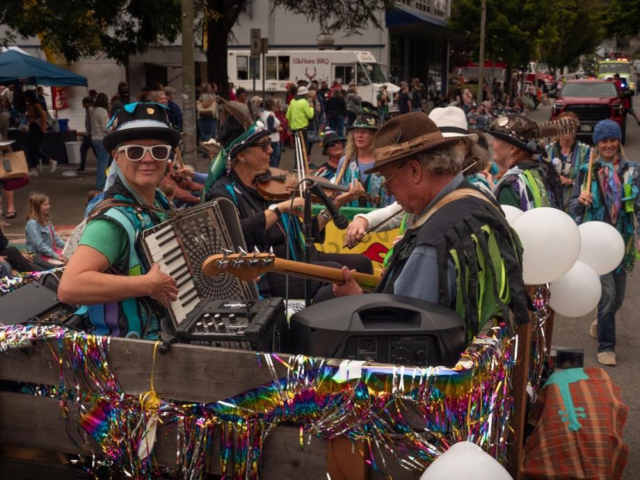 Colorfully costumed musicians playing accordion, violin, and guitar perform on a festive, tinsel-decorated float during the Bay Area Fun Festival parade in Coos Bay, Oregon, with crowds lining the street in the background.