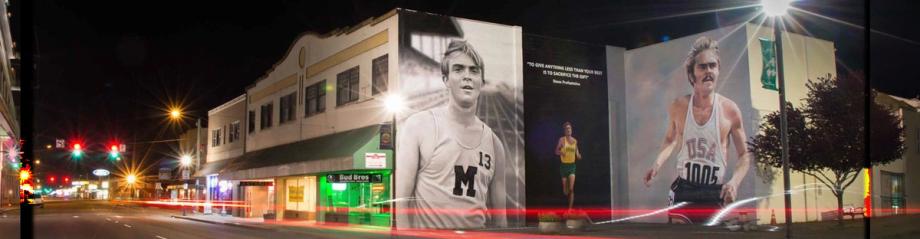 A large mural of runner Steve Prefontaine lit up at night in downtown Coos Bay, featuring his quote 'To give anything less than your best is to sacrifice the gift.'