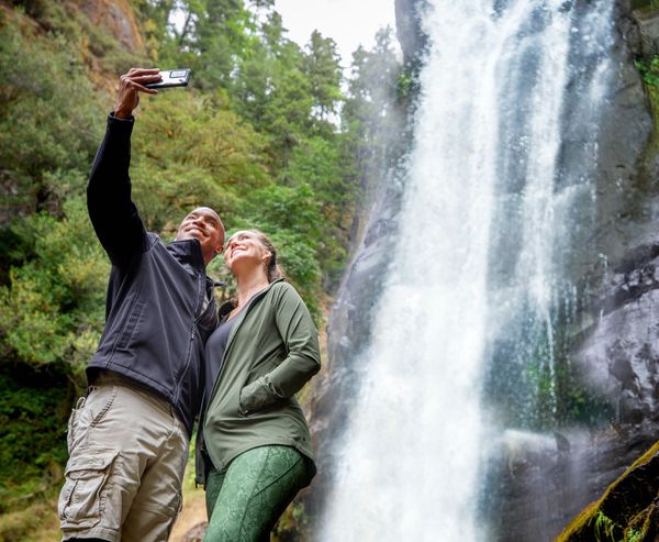 A couple takes a selfie together in front of a tall waterfall cascading down a rocky cliff, surrounded by forest.