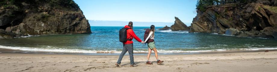 alt="A couple with backpacks walks hand-in-hand along Simpson Beach, framed by dramatic rocky headlands and bright blue ocean waters on a clear day.