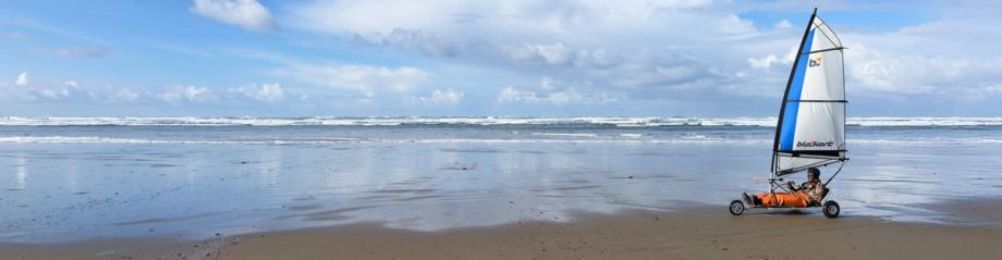 A person rides a wind-powered land sailing cart with a blue and white sail across the wet sand at Bastendorff Beach, with ocean waves in the background.