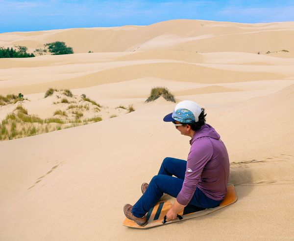 A sandboarder sits atop a board, poised to ride down a large dune surrounded by rolling sand hills.