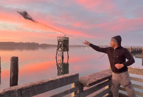 A man throwing a crab ring net from a pier at Hollering Place under a vivid pink and orange sunrise sky reflected in the calm water below.