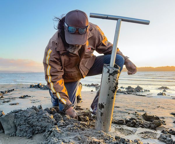 A clammer uses a clam gun to dig in the wet sand on an Oregon Coast beach at sunrise, kneeling close to the ground to retrieve clams by hand.