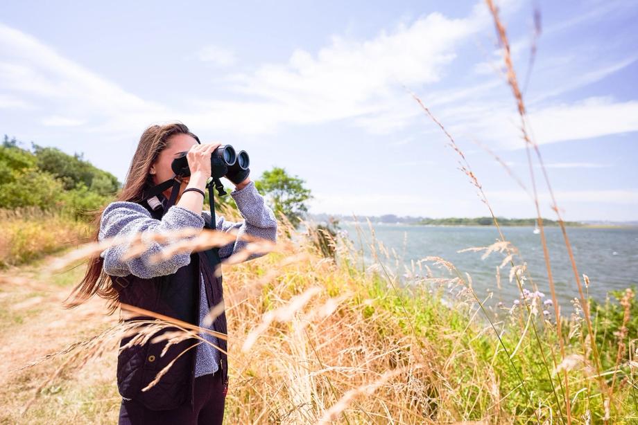 A young woman looks through binoculars along a coastal trail of tall grasses, with the bay and distant shoreline behind her.
