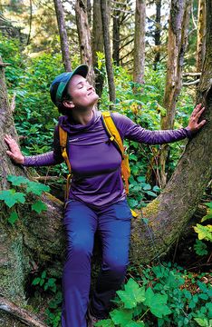 Woman in hiking gear leaning against mossy trees with eyes closed, forest bathing among lush green Pacific Northwest woodland.