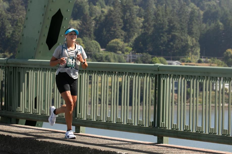 A female runner wearing a race bib, blue visor, and hydration vest crosses a green metal bridge over the bay during the Circle the Bay 30K race, with forested hills visible in the background