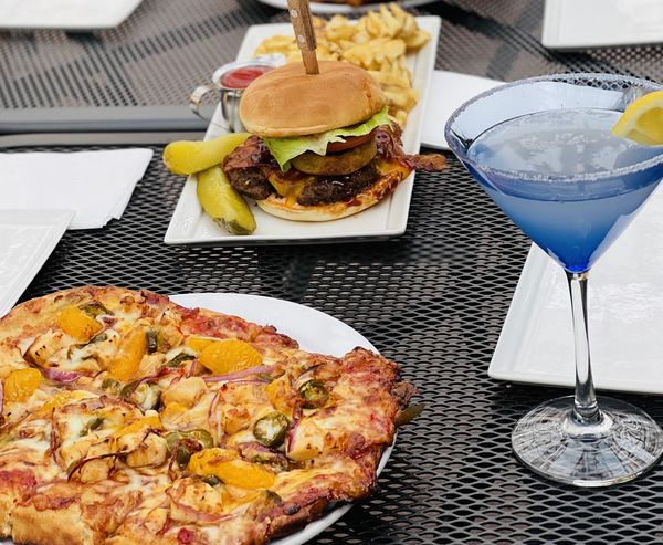 Bacon cheeseburger with fries, jalapeño mandarin pizza, and a blue cocktail on an outdoor patio table.