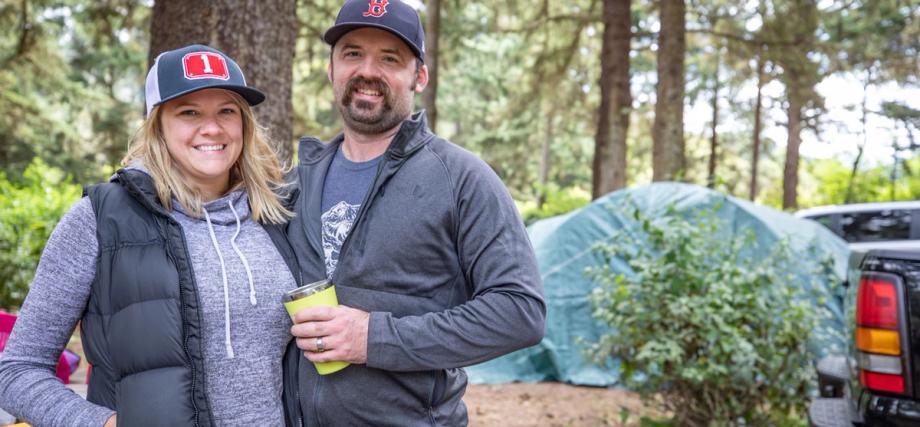 A smiling couple poses at a forested campsite at Sunset Bay State Park, with a tent and vehicle visible in the background.