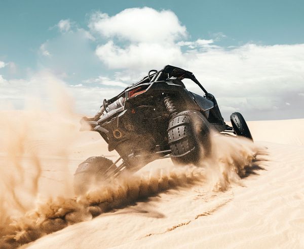 A dune buggy catches air while carving down a sandy slope, kicking up a large spray of sand.