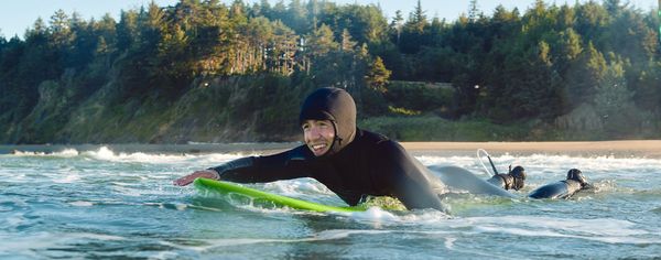 Surfer in a wetsuit paddling out through a wave near a forested coastline.