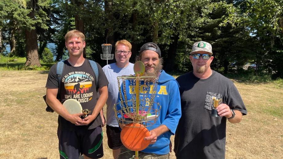 Four men posing outdoors with a disc golf basket holding trophies and discs, standing on grass in front of tall evergreen trees.