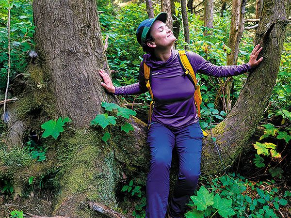 A woman closes her eyes and presses her hands against two mossy tree trunks, practicing forest bathing and mindfulness in the lush forests of Oregon's Adventure Coast.