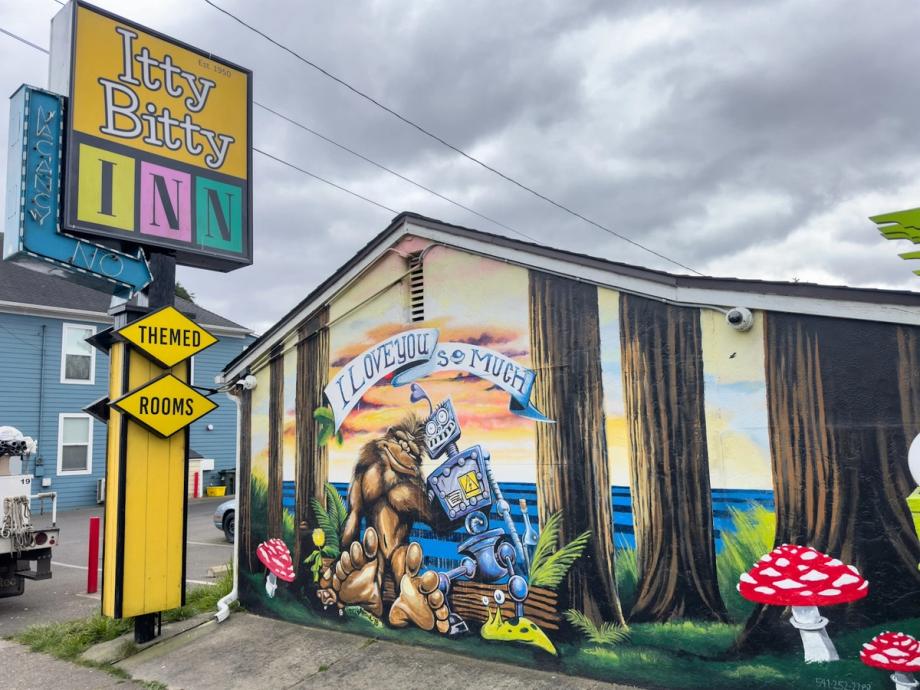 Retro yellow, pink, and teal Itty Bitty Inn motel sign advertising themed rooms, next to a colorful mural of Bigfoot embracing a robot under a banner reading I Love You So Much, surrounded by painted trees and red-and-white mushrooms, under an overcast sky.