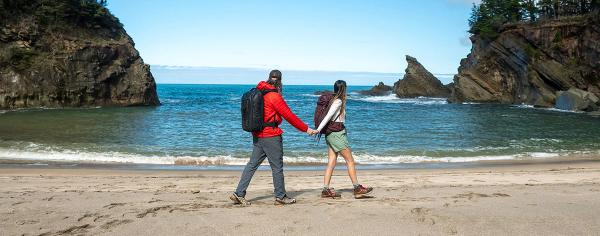 A couple walks hand in hand along Simpson Beach, with dramatic rock formations flanking a bright blue cove on Oregon's Adventure Coast.