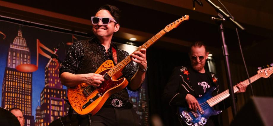 Smiling guitarist in white sunglasses and a patterned shirt plays an ornate orange Telecaster on stage, with a bassist in the background.
