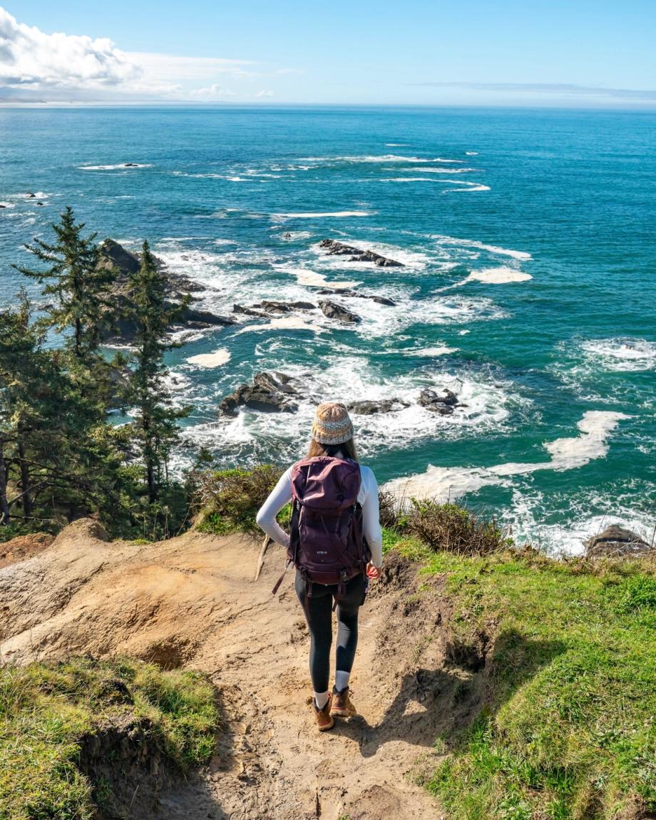 Person with backpack standing on a cliffside trail above rocky shoreline on a Sunny Day at Cape Arago State Park, Coos County, Oregon.